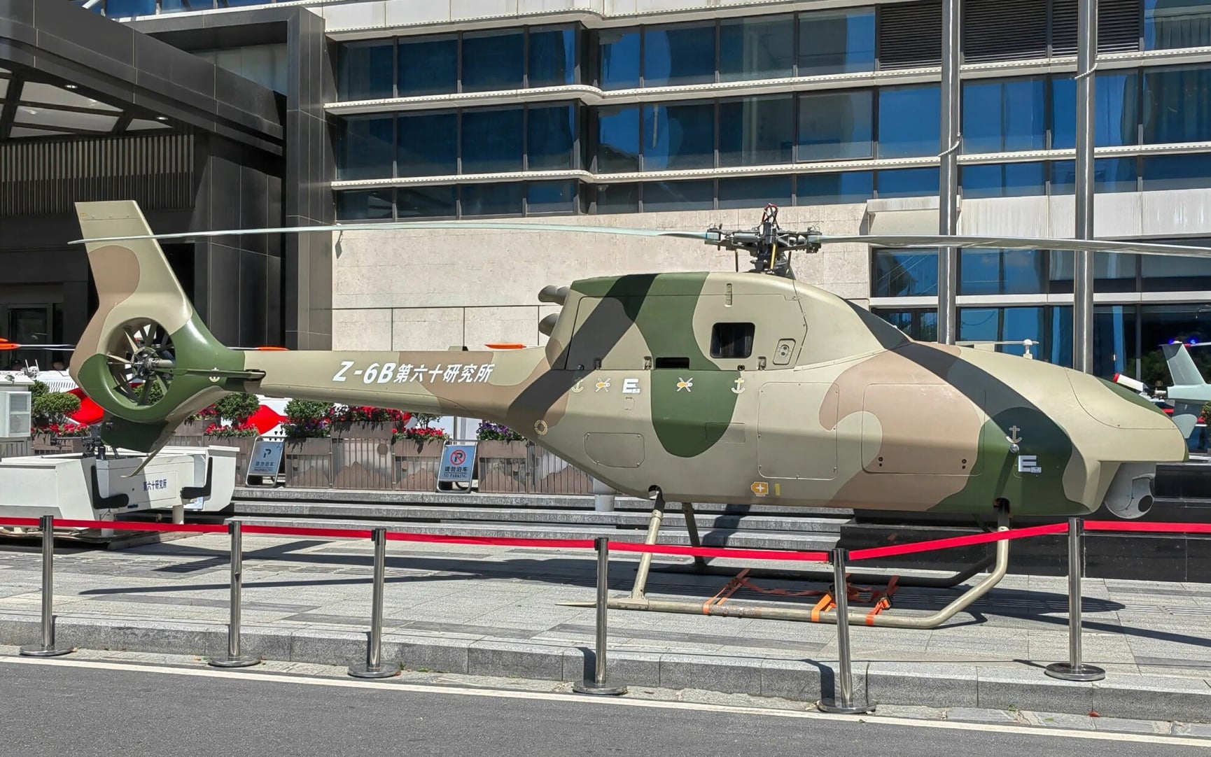 Camouflaged Z-6B unmanned helicopter roped off outside a glass office building.