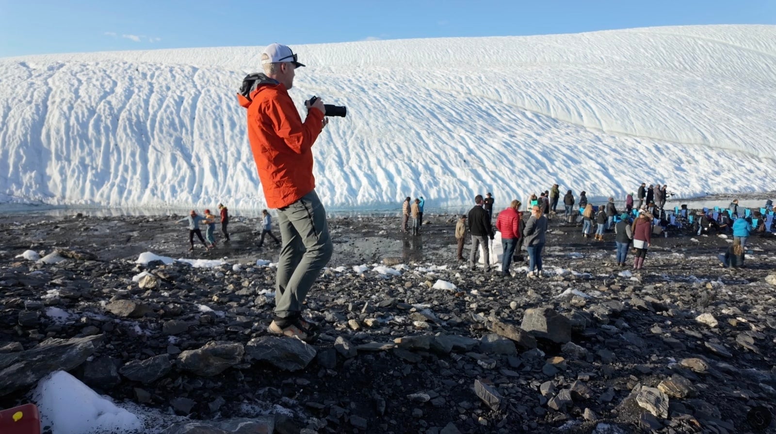 Videographer in an orange jacket filming the crowd gathered below an Alaskan glacier.