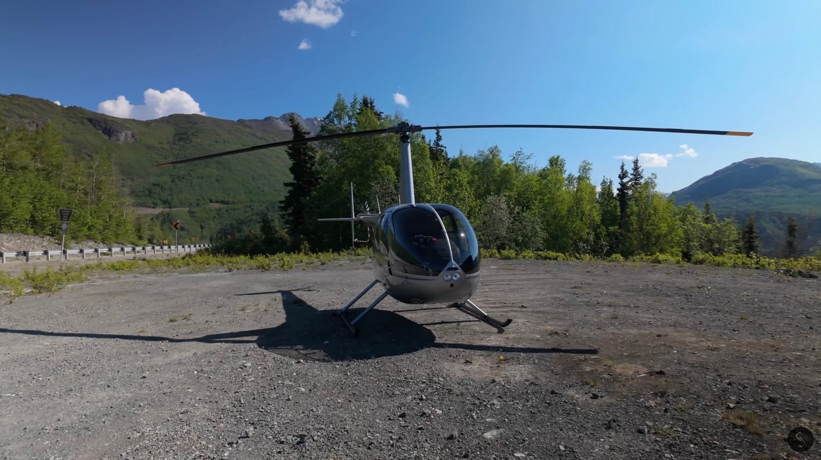 Small helicopter parked on a gravel pad ringed by spruce forest and mountains.