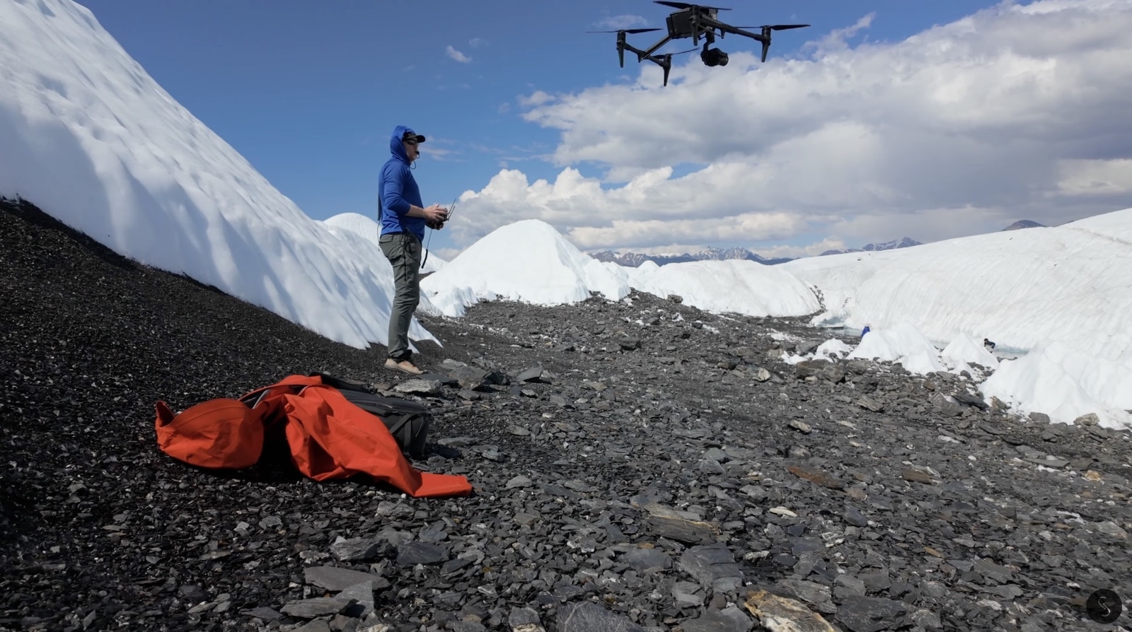 A DJI Inspire 3 flying above a glacier moraine as its pilot watches.