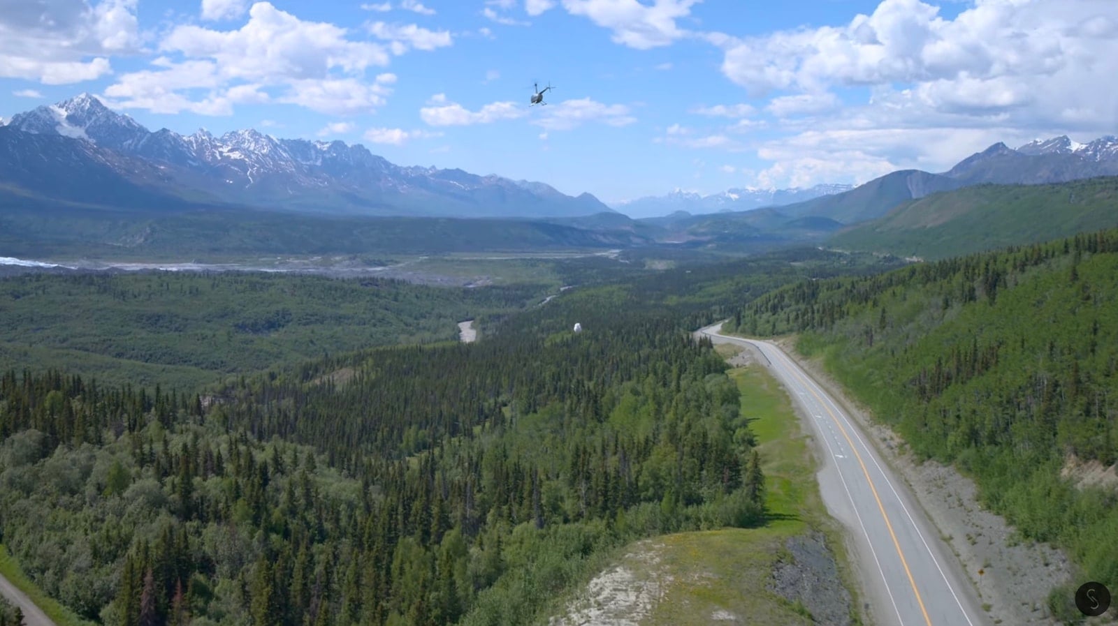 Helicopter flying above a highway cutting through spruce forest and snow-capped peaks.