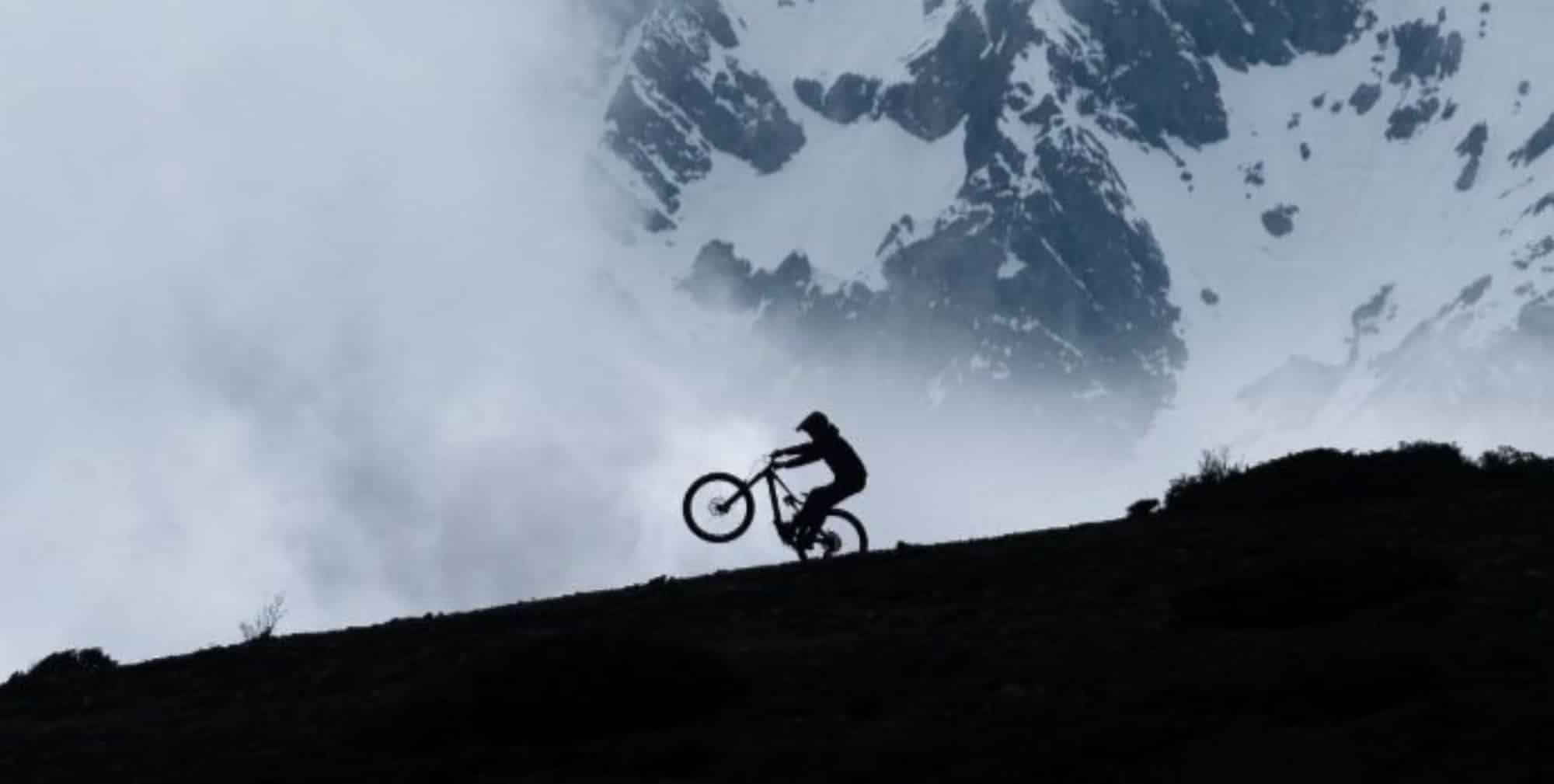 A rider silhouetted on a ridgeline with snowy peaks and cloud behind.