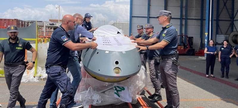 Guardia di Finanza officers handling a large wrapped drone fuselage section inside a hangar.