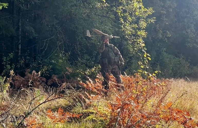 Camouflaged soldier hand-launches a small fixed-wing reconnaissance drone from a sunlit forest clearing.