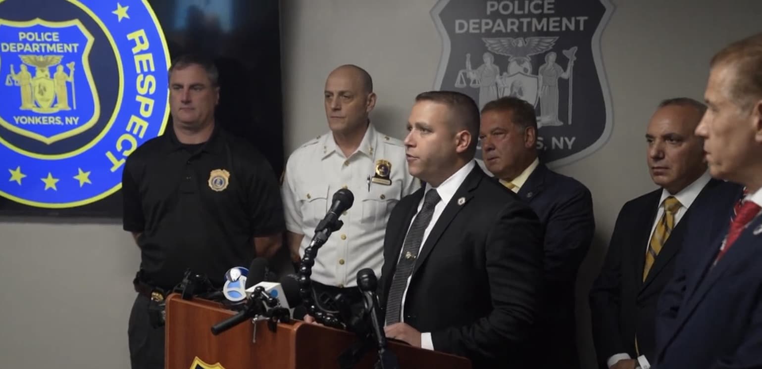 Officials in suits and uniforms stand at a Yonkers Police Department podium.