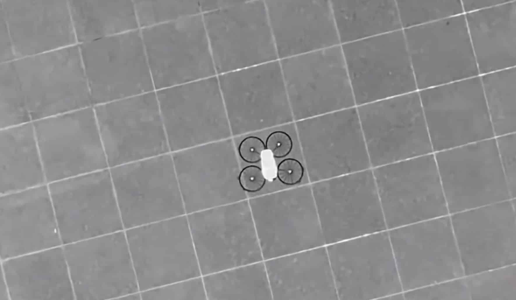 Small white quadcopter hovers above a tiled plaza in a black and white aerial frame.