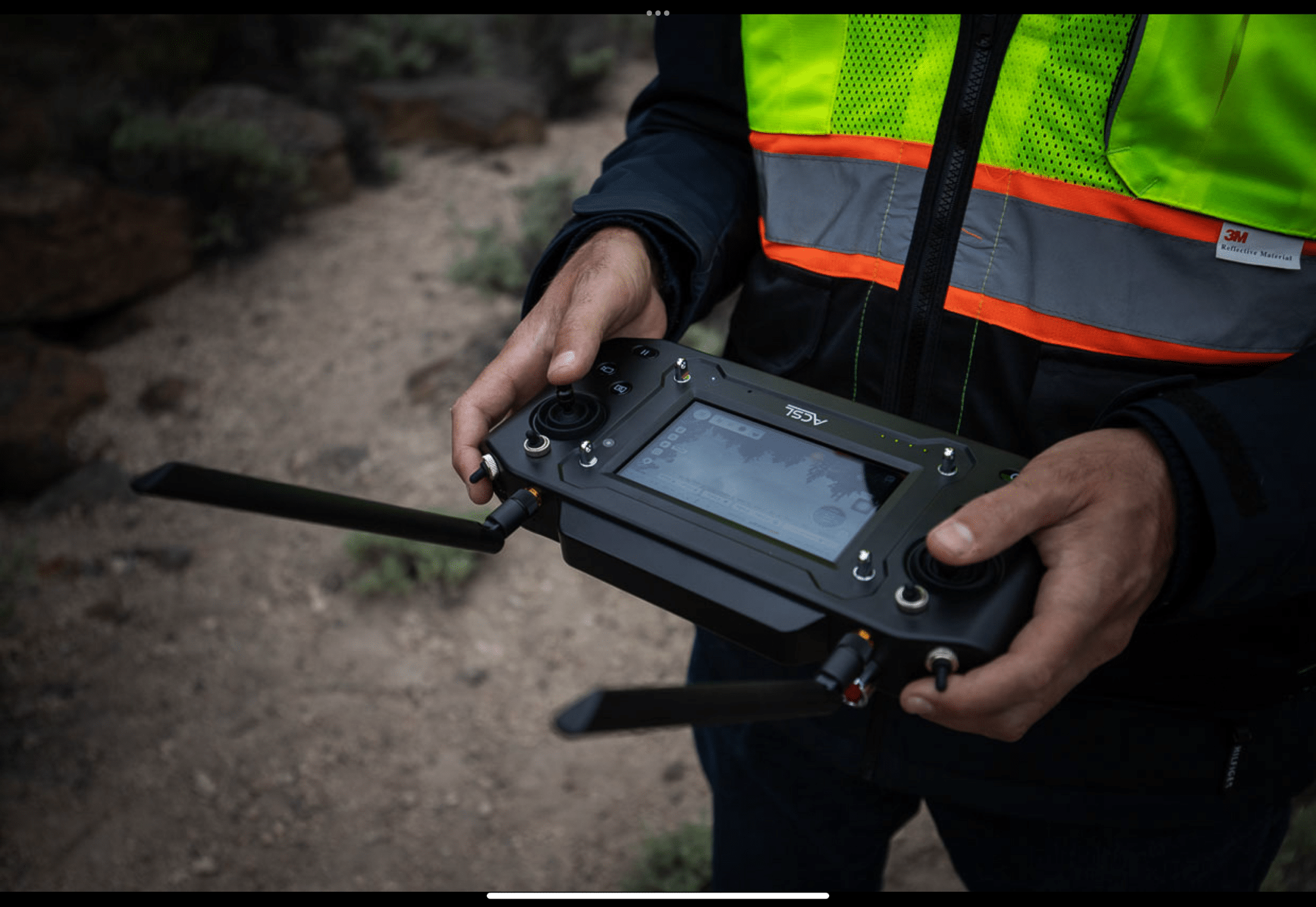 Operator in a hi-vis vest holding the ACSL TENSO smart controller in desert scrub.