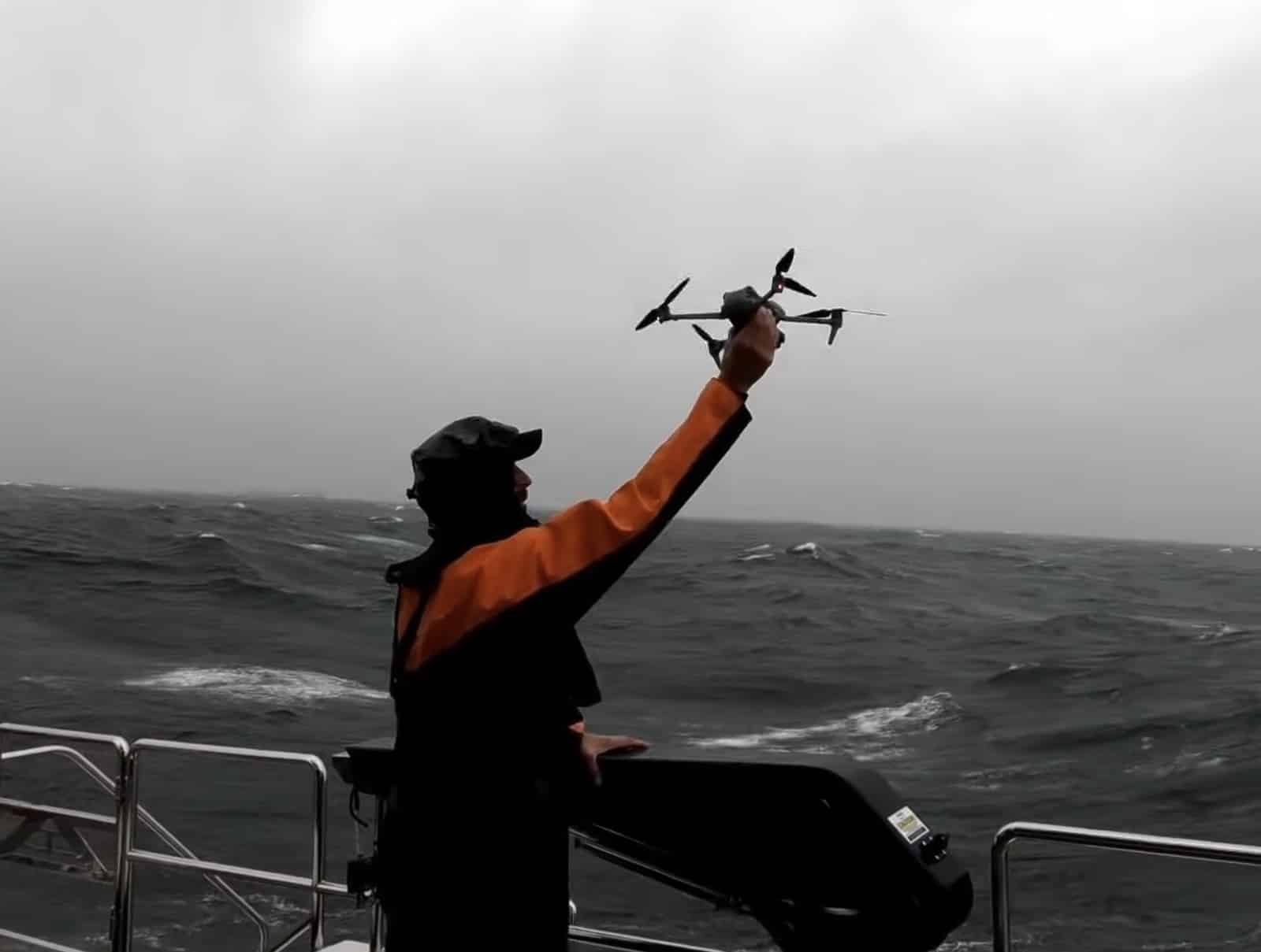 Crew member in orange foul-weather gear hand-catching the drone on a pitching deck.