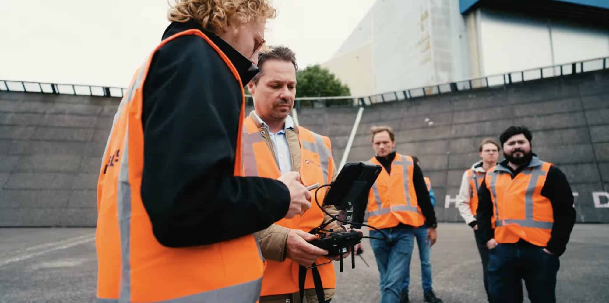 Crew in orange vests flying an inspection drone beside a Schiphol maintenance building.