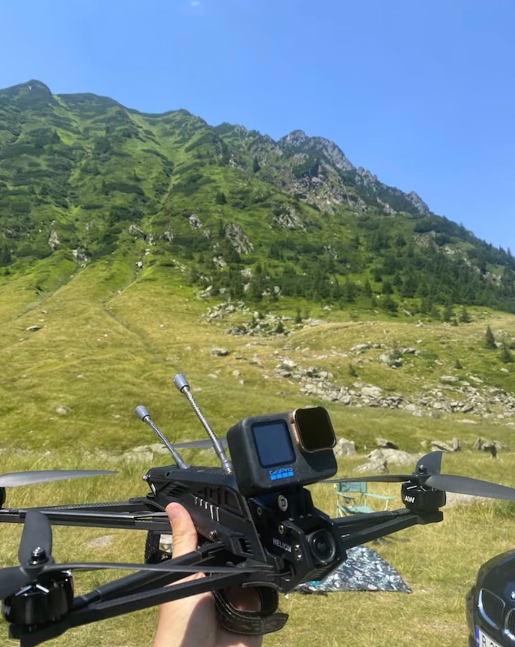 Hand holding an FPV quad with a GoPro on top on a green mountain slope.