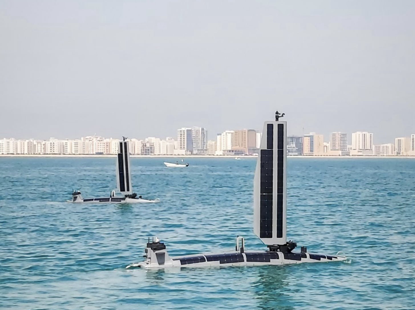 Two solar-sail unmanned surface vessels underway off a coastal city skyline.