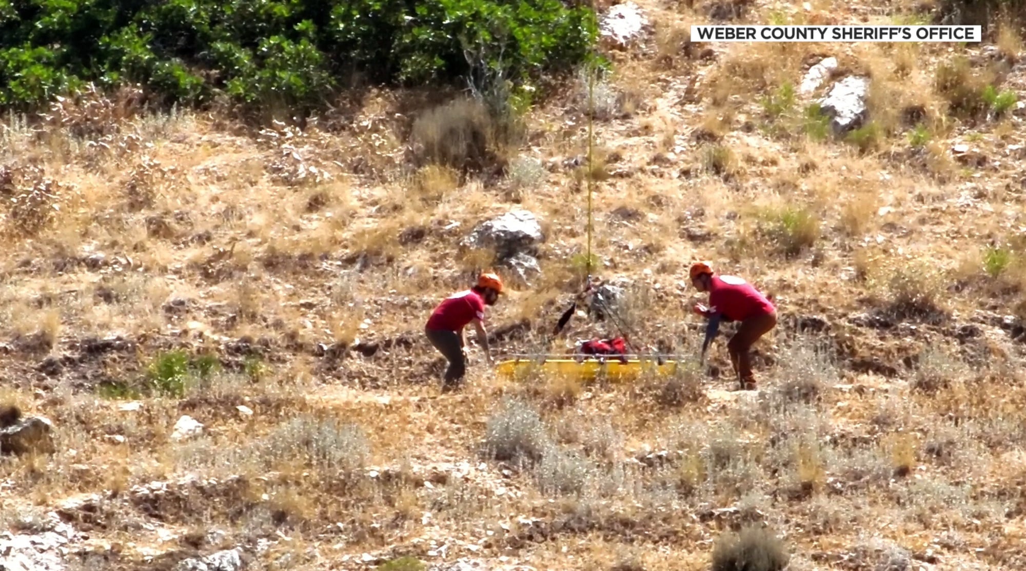 Two rescuers in red shirts receive a yellow litter lowered by winch line uphill.