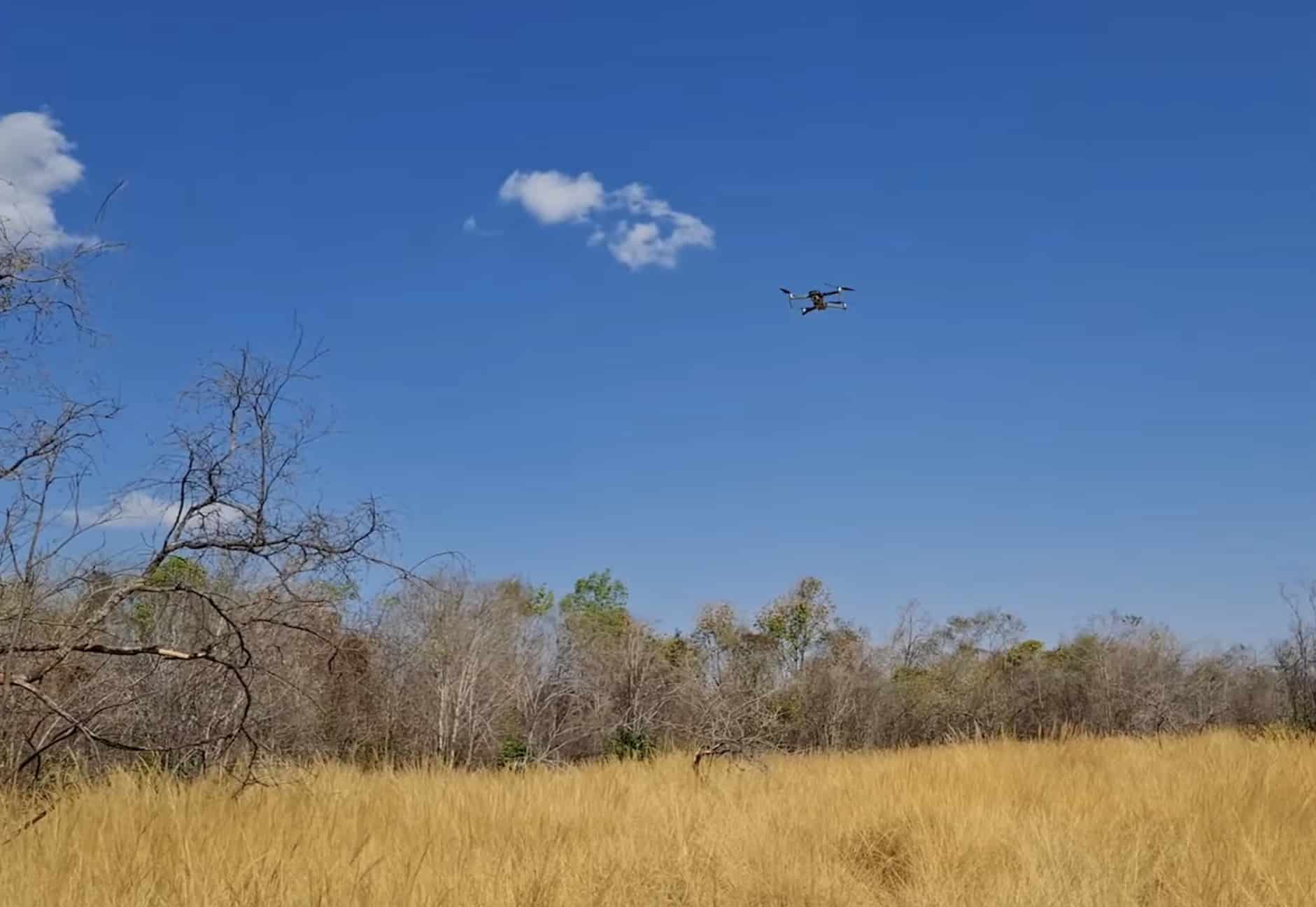 Small drone hovering high above dry grassland and scrub under a blue sky.