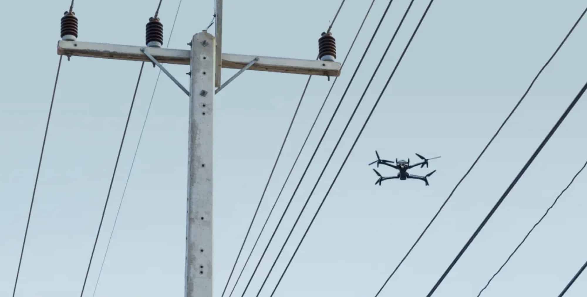 Black inspection quadcopter hovers beside a concrete utility pole and power lines.