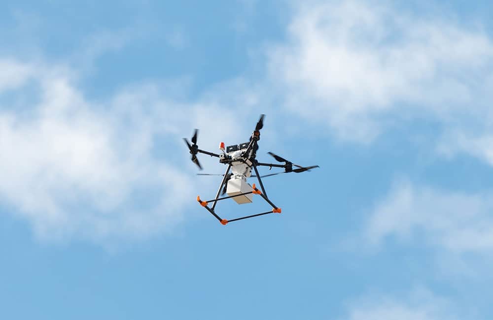Delivery drone carrying a white parcel across a bright blue sky.