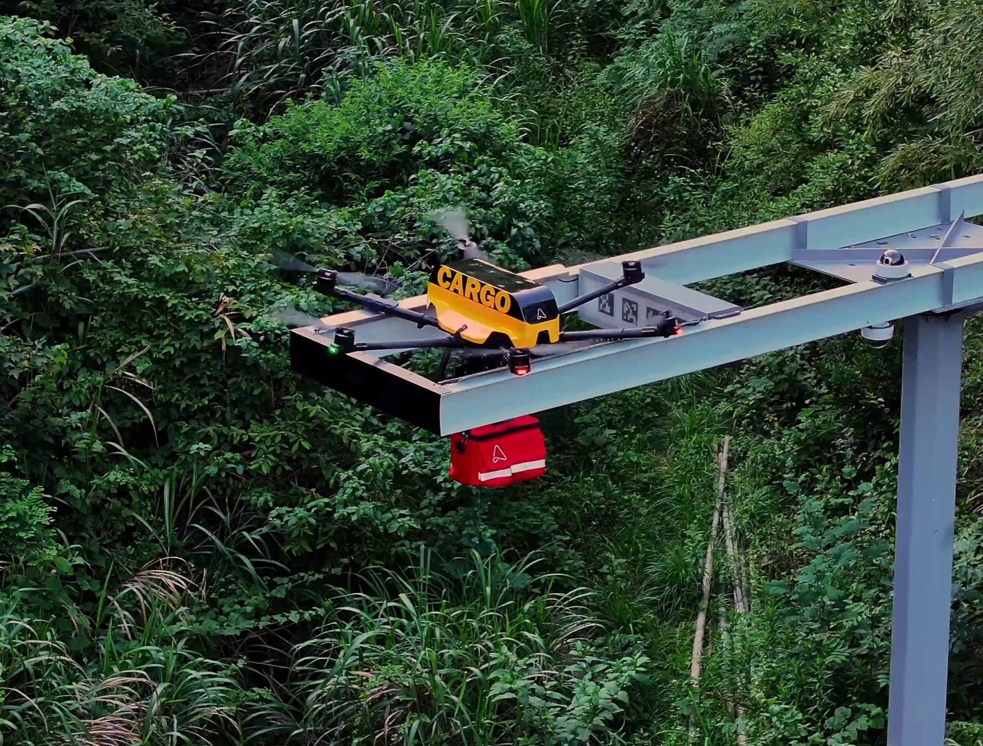 Yellow cargo quadcopter settling onto an elevated steel dock above dense jungle.