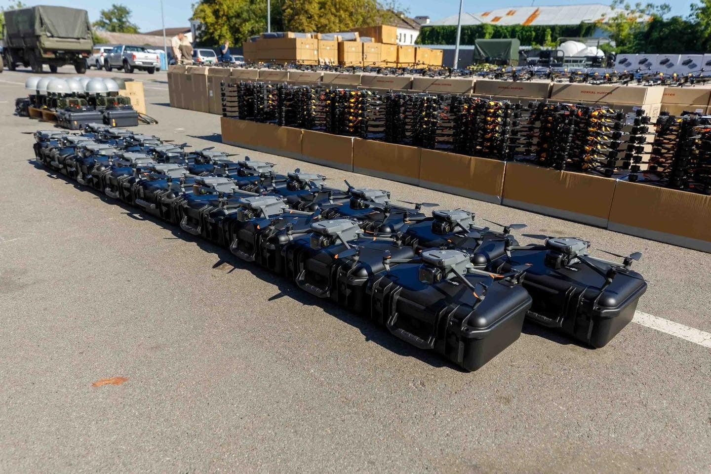 Rows of hard cases holding DJI Mavic drones on tarmac beside military trucks.