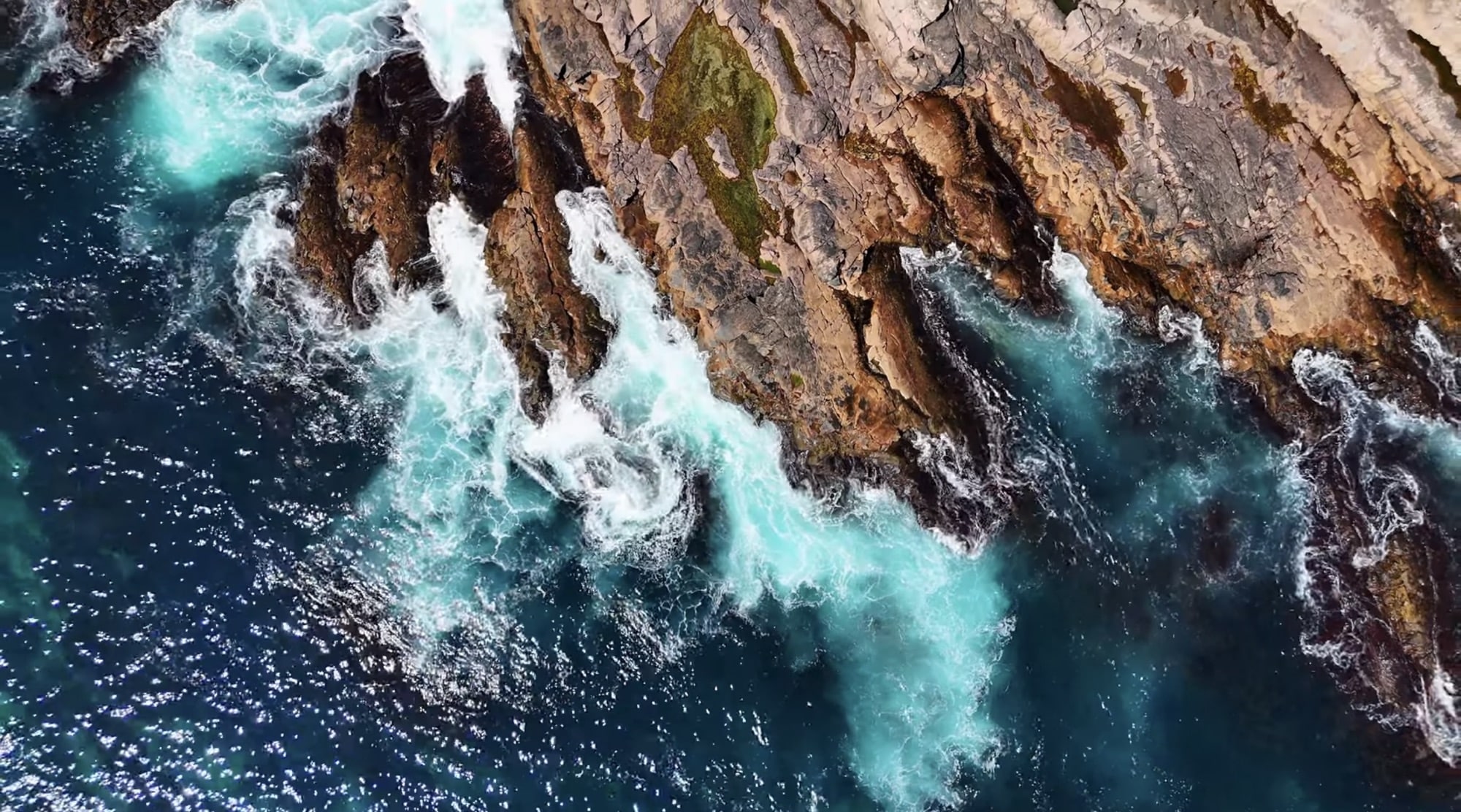 Top-down aerial of turquoise surf breaking against a rugged, weathered rock shelf.