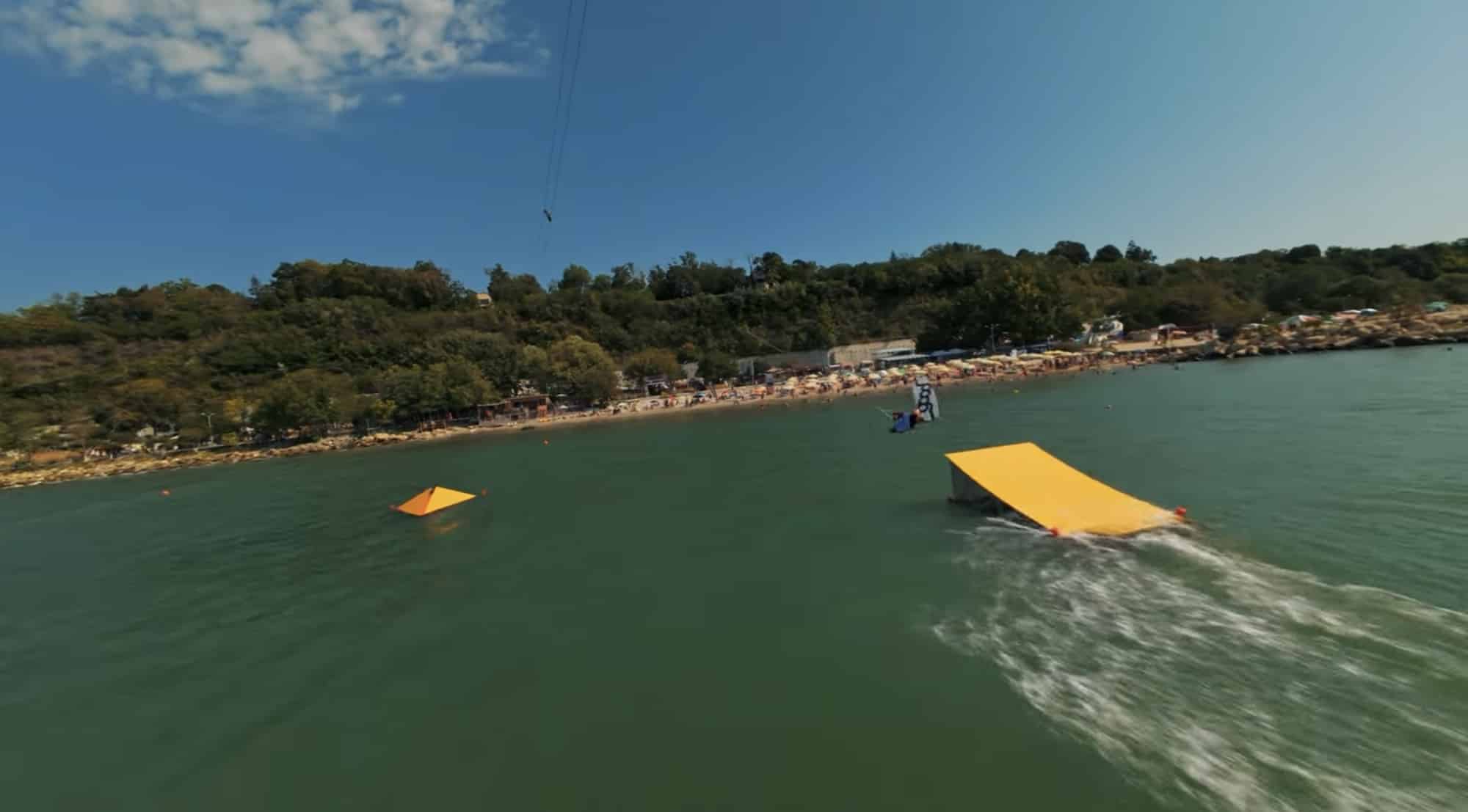 FPV view over a cable wake park with yellow ramps and a rider below.