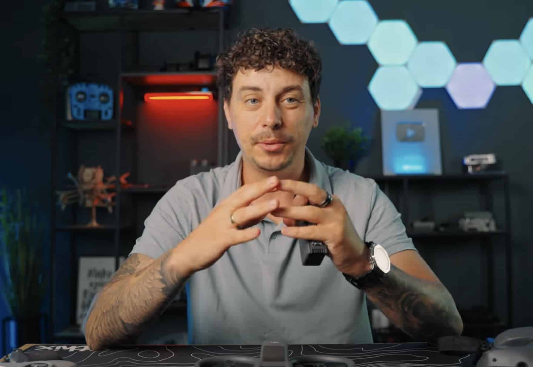 Reviewer talking to camera in a studio lit by hexagon panels and drone shelves.