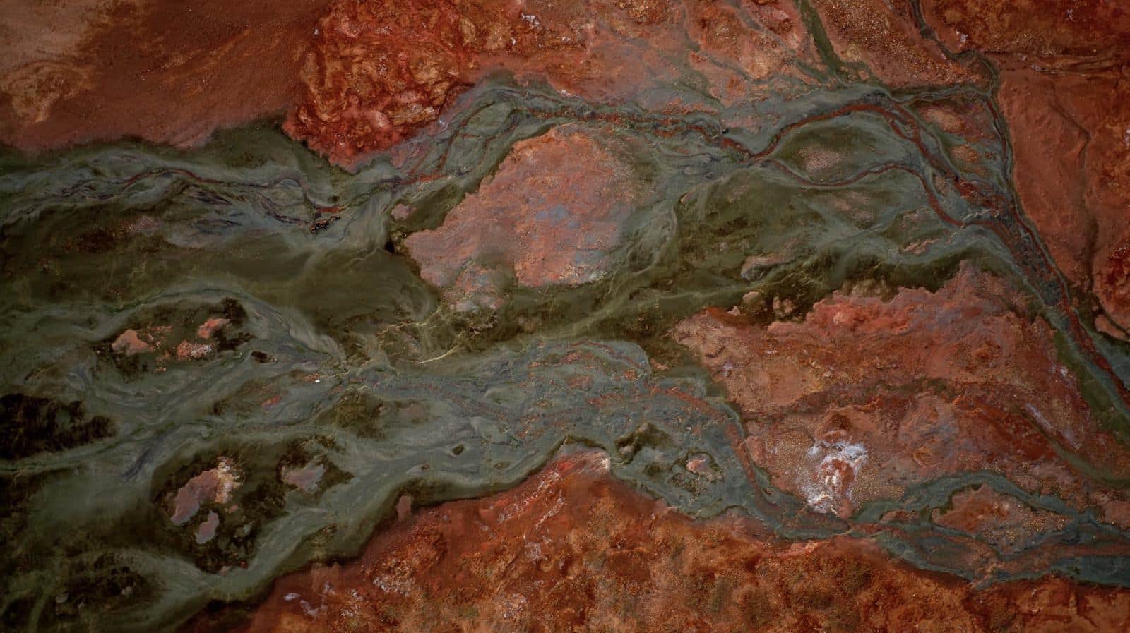 Abstract aerial of rust-red and green mineral flats cut by braided meltwater channels.