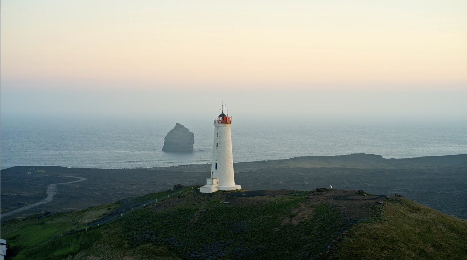 White lighthouse on a headland at dusk with a sea stack offshore.