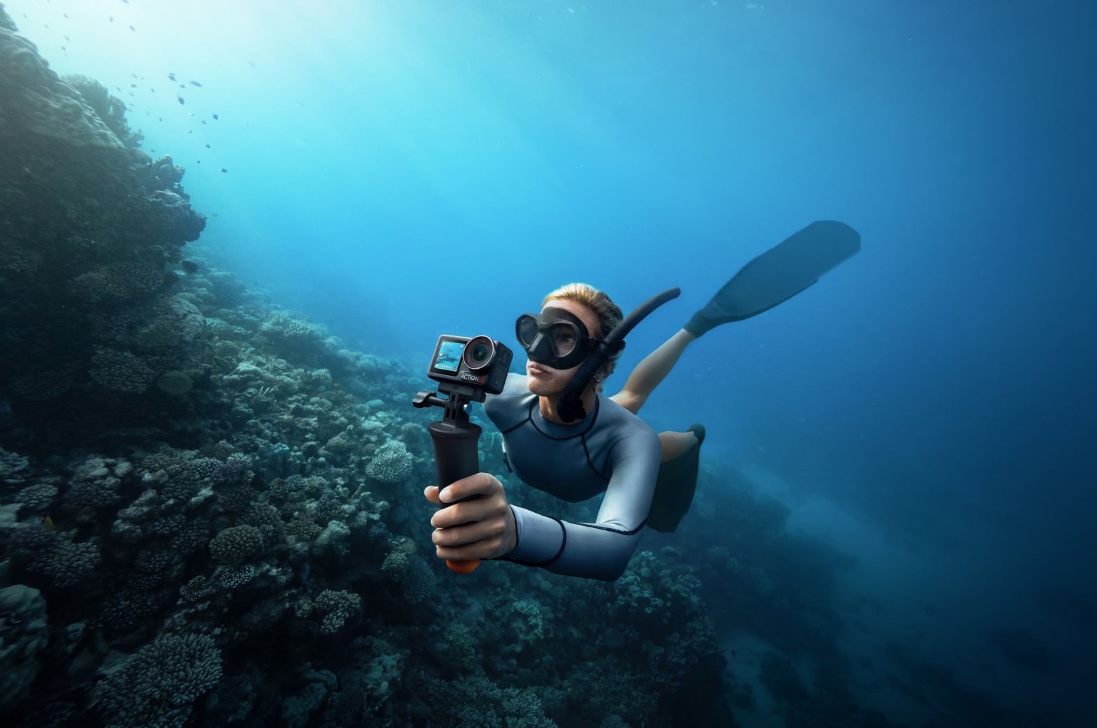 Freediver holding the camera on a grip while swimming along a coral wall.