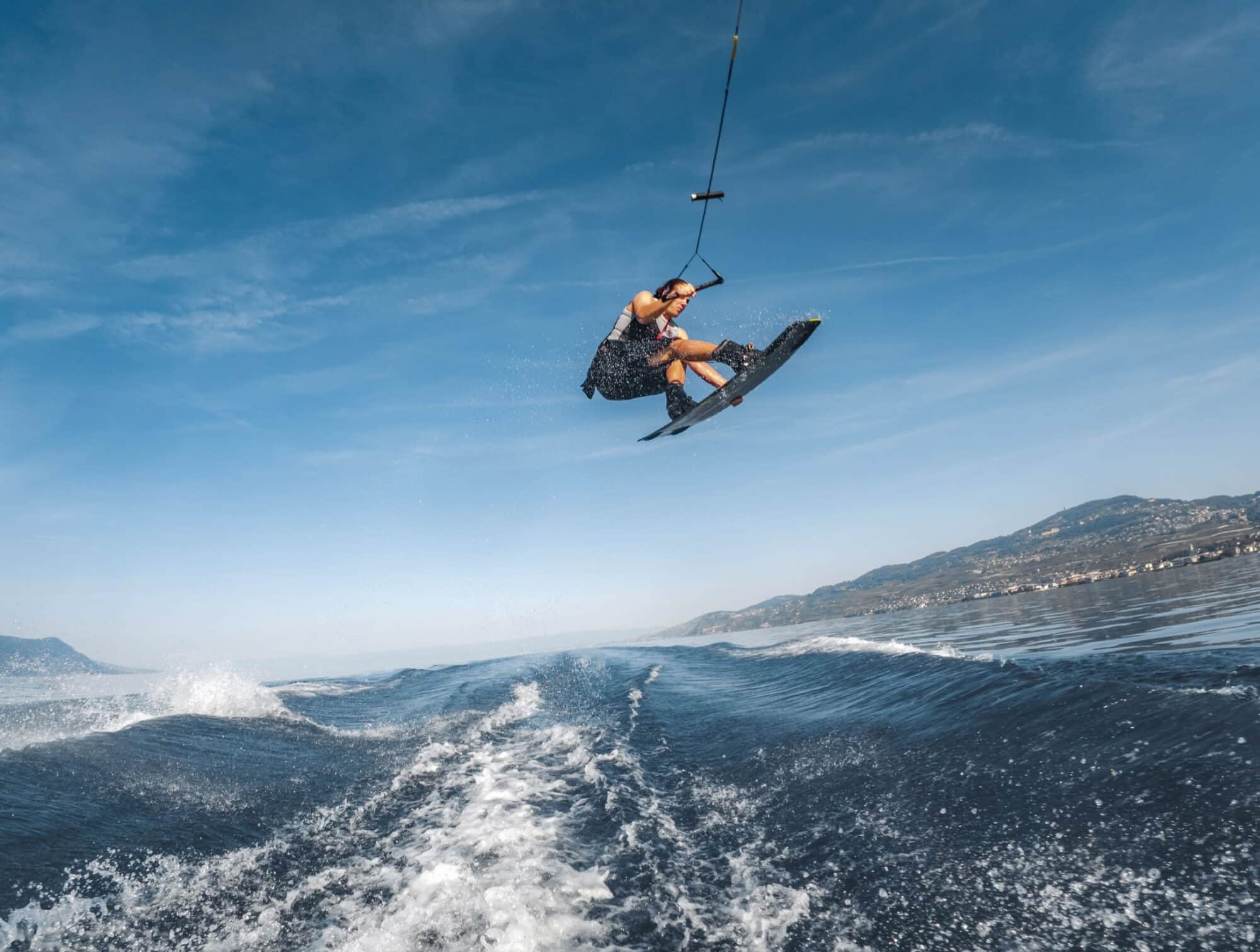 Wakeboarder airborne above the boat wake with the tow handle stretched overhead.