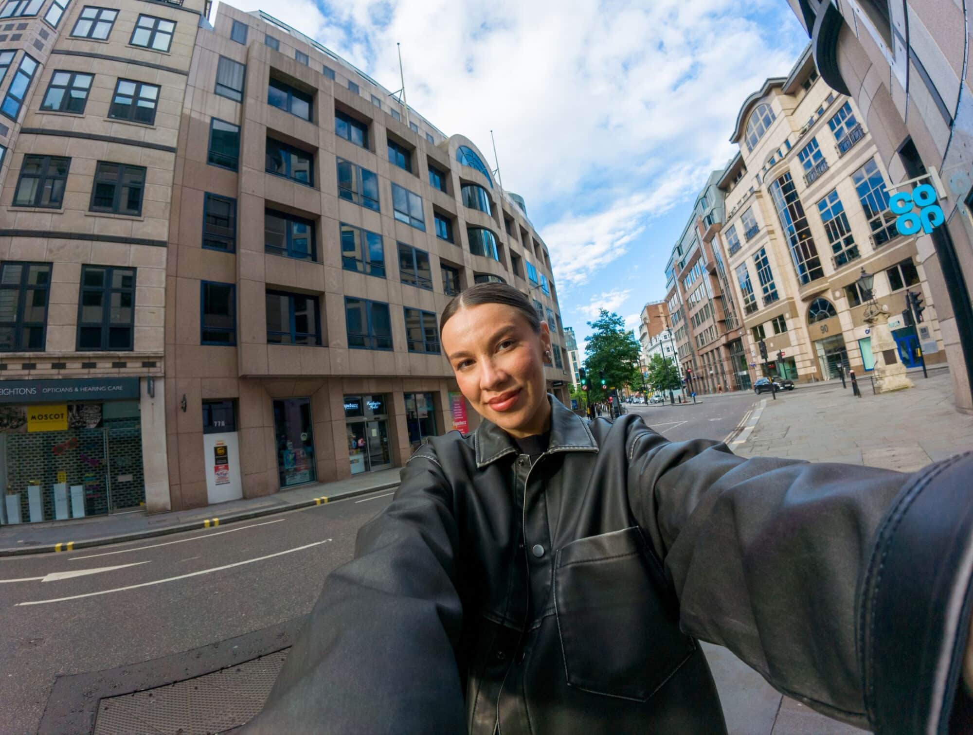 Selfie-style wide shot of a woman in a leather jacket on an empty street.