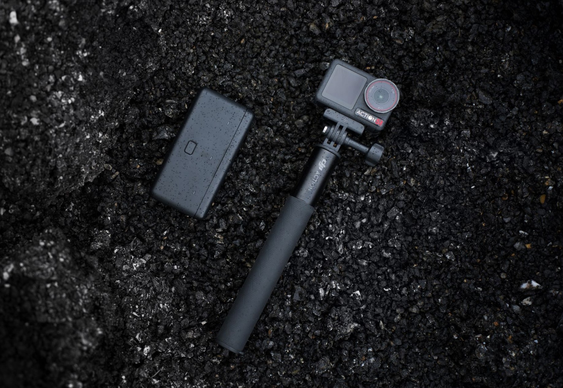 Camera on a mini extension rod beside a battery case on dark gravel.
