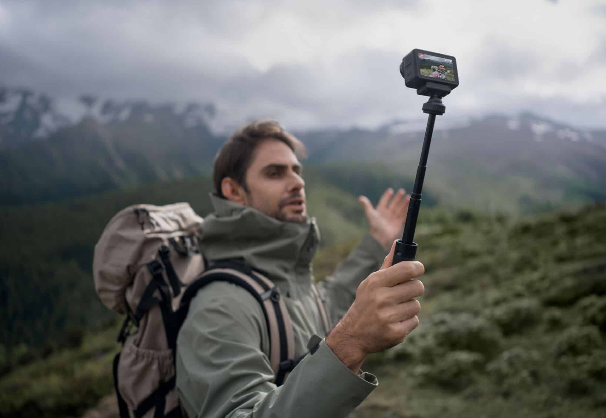 Backpacker holding the camera on an extension rod in front of snowy mountains.