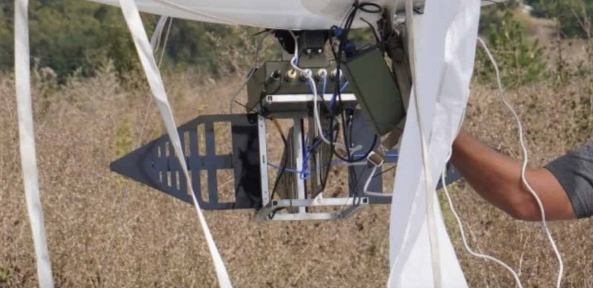 Electronics pod suspended beneath a white aerostat envelope as a hand adjusts cables.