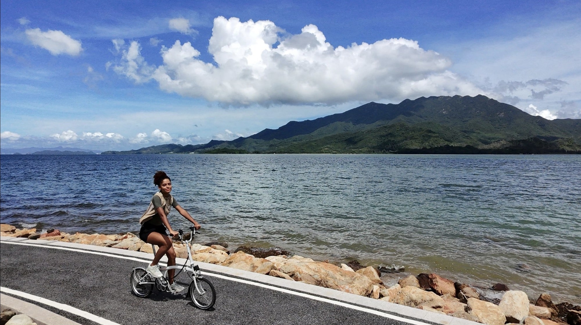 Neo footage of a cyclist on a lakeside road with mountains beyond.