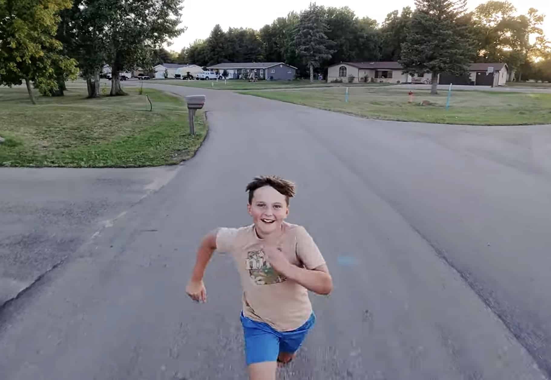 Drone view of a grinning boy running toward the camera down a rural driveway.