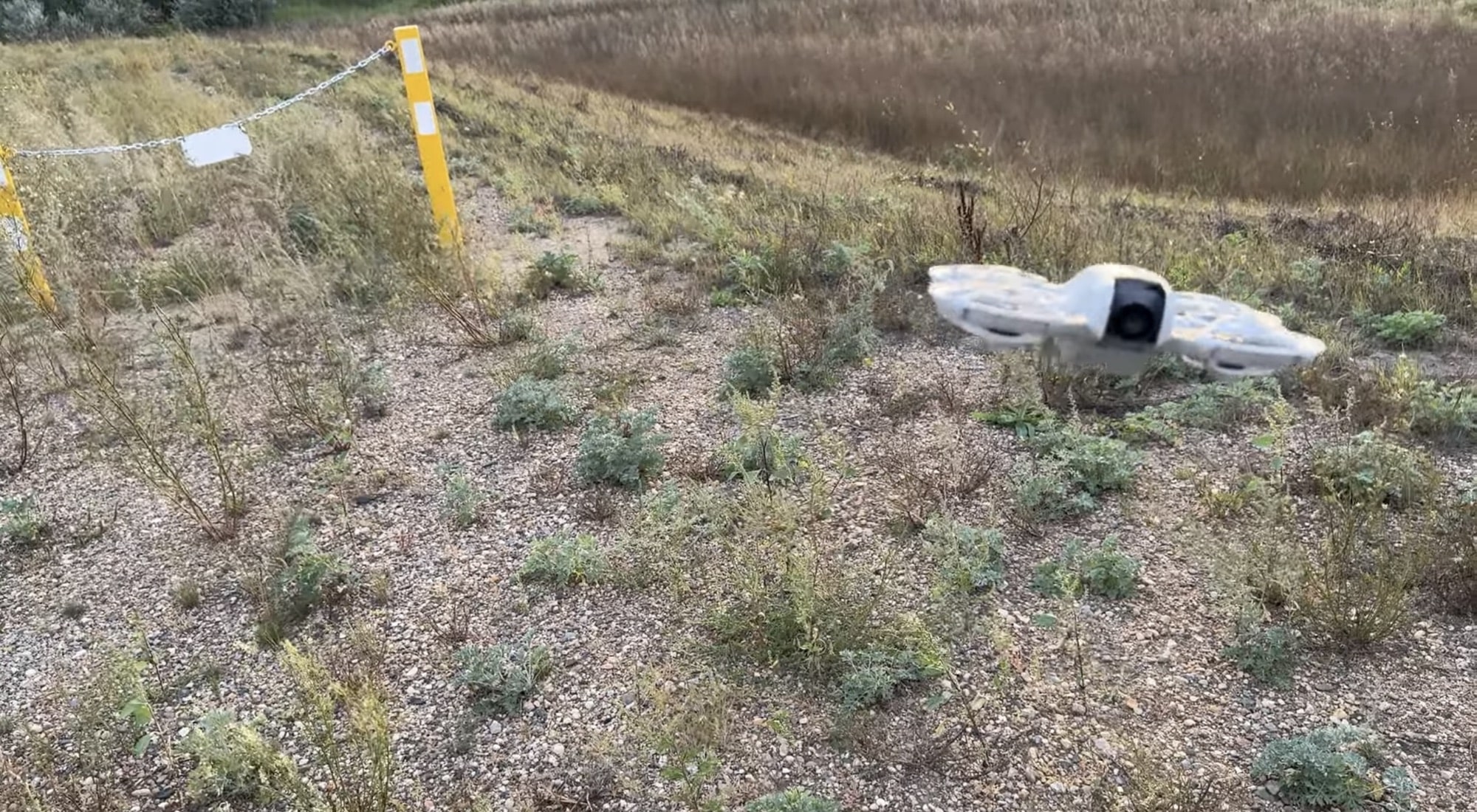 DJI Neo hovering over dry weeds beside a striped roadside marker post.