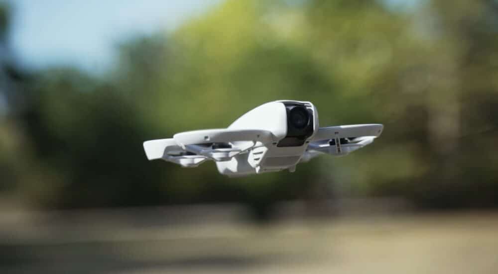 DJI Neo hovering with its propeller guards removed against blurred green trees.