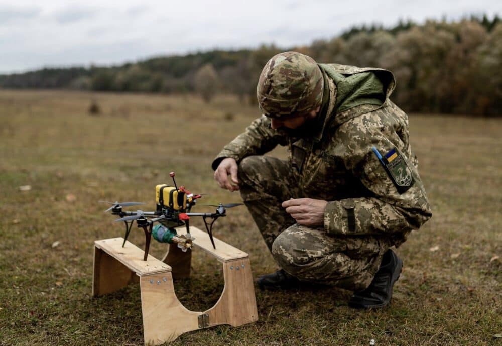 Ukrainian soldier crouches beside an FPV quadcopter carrying a payload on a plywood stand.