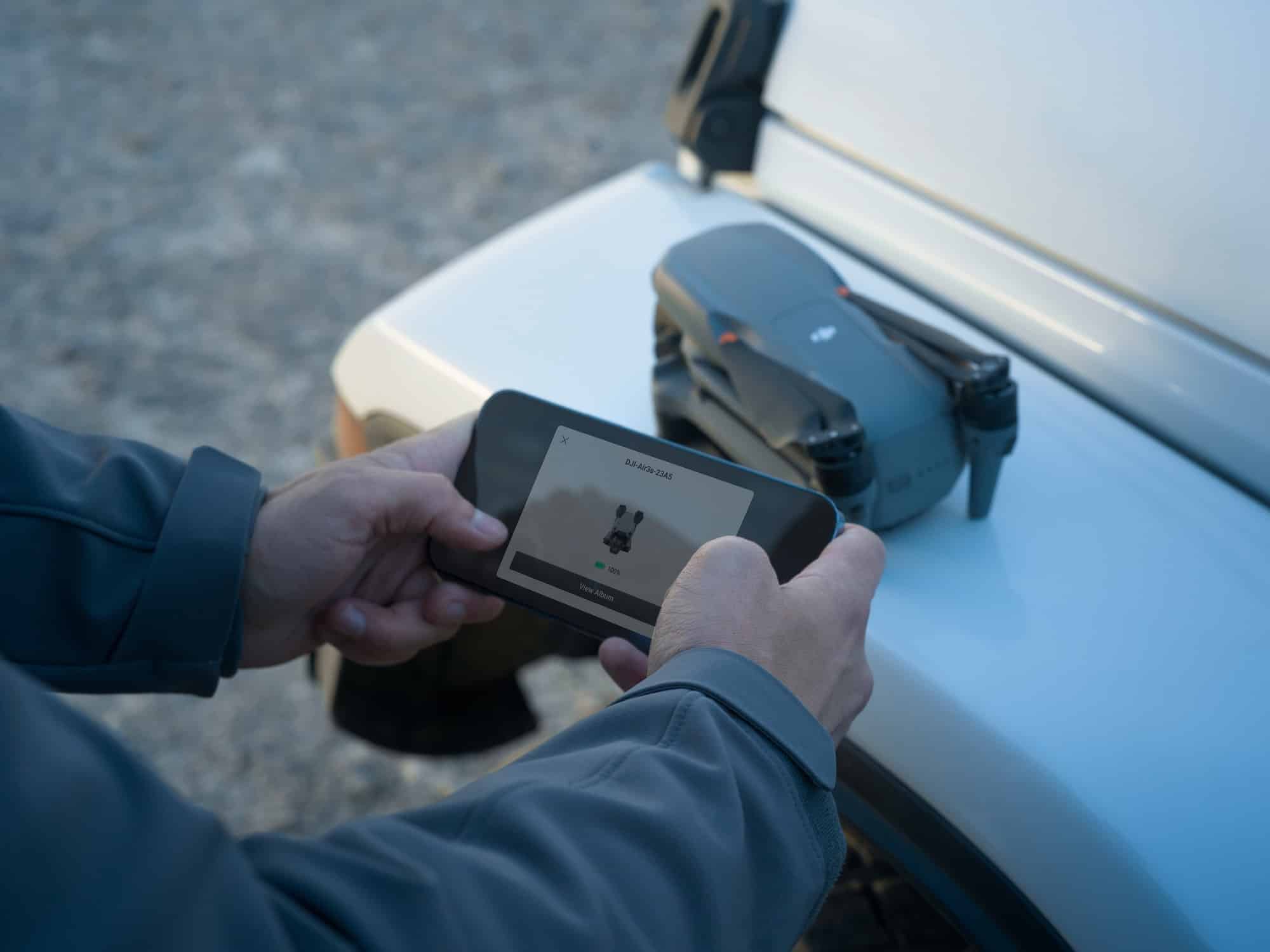 Hands hold the RC 2 controller over a white car hood with the drone alongside.
