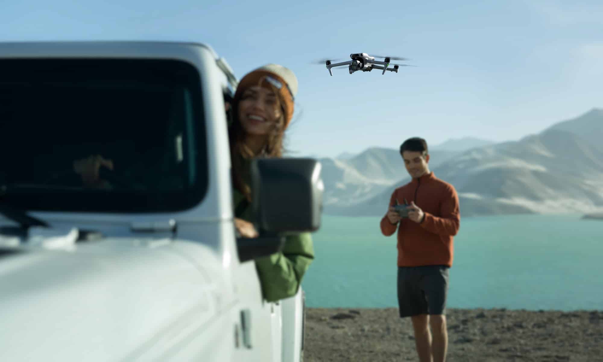 Woman leans from a white pickup with a controller as the drone hovers nearby.