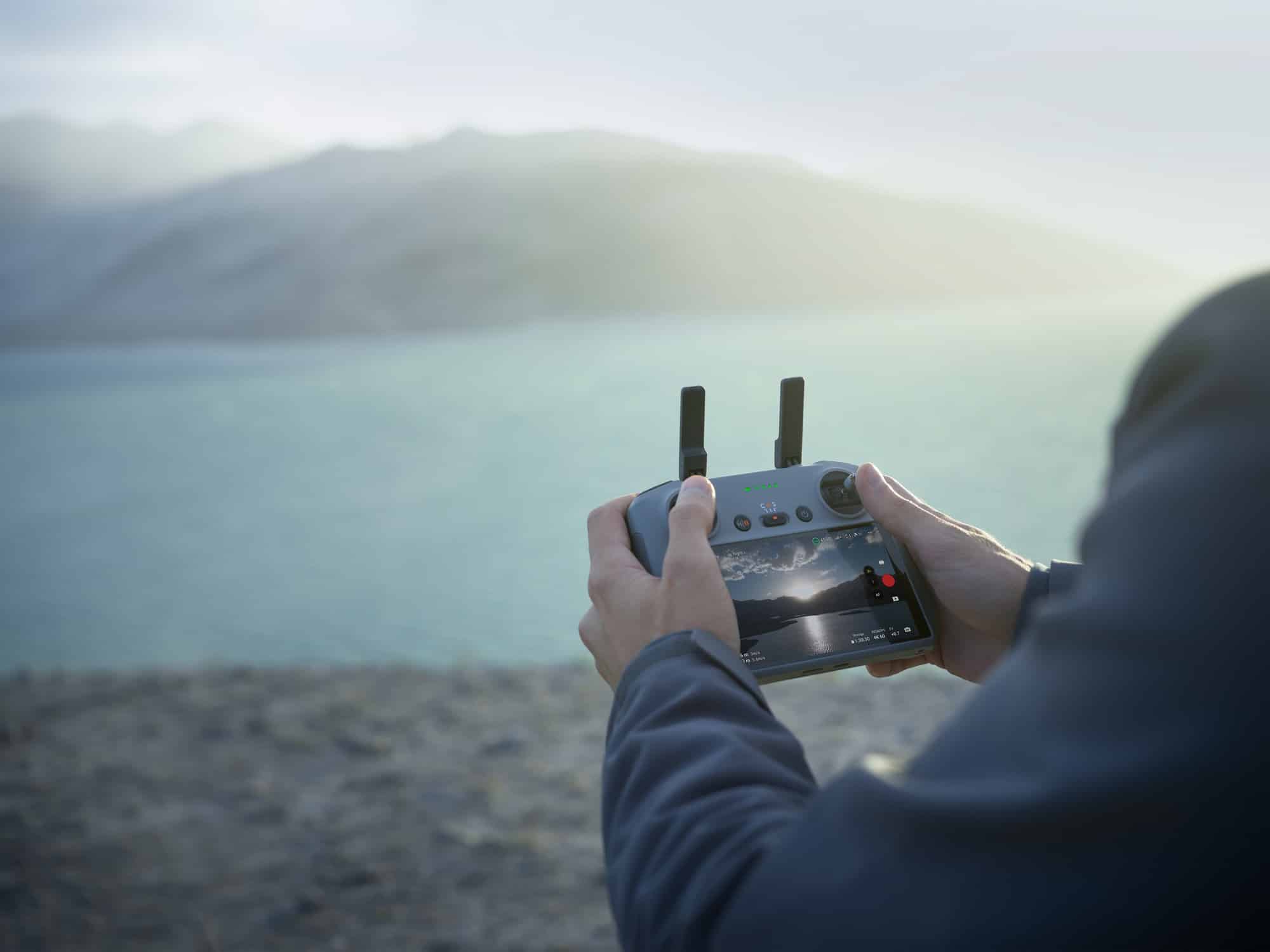 Hands grip the DJI RC 2 controller with a hazy lake and mountains behind.