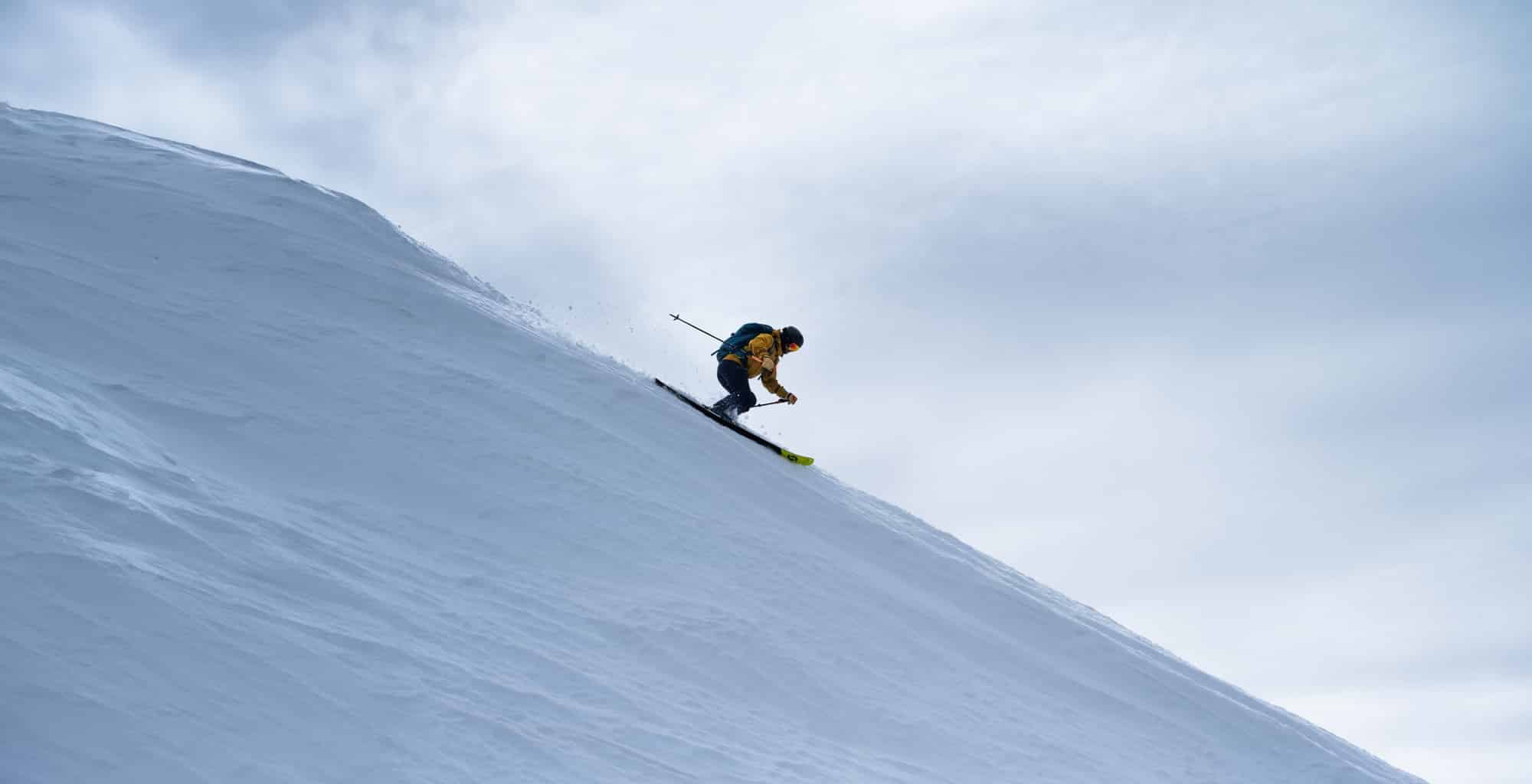 Lone skier descends a vast unbroken snow face in a distant wide shot.