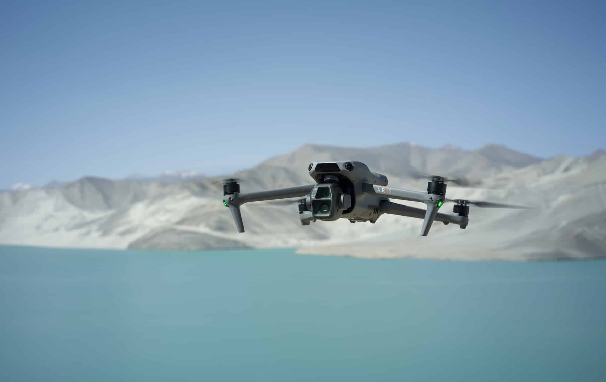 Air 3S flying head-on across a pale turquoise lake ringed by bare mountains.