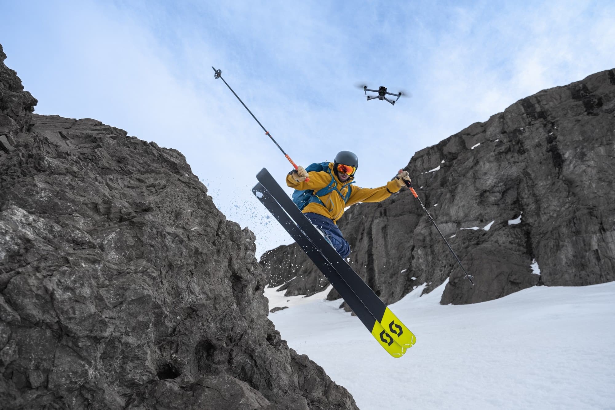 Skier jumps a rock couloir with the drone visible high in the sky.