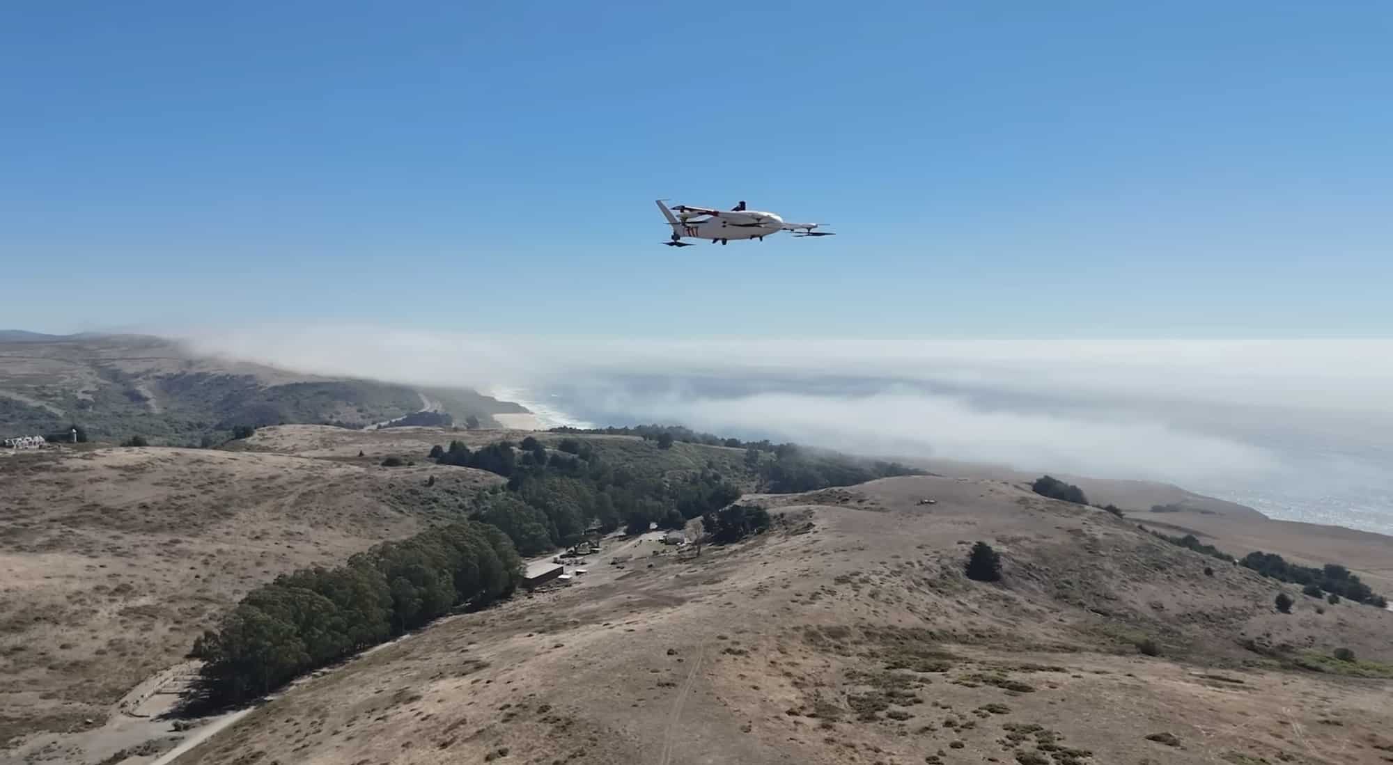 Zipline delivery aircraft flies above coastal hills with fog rolling over the ocean below.