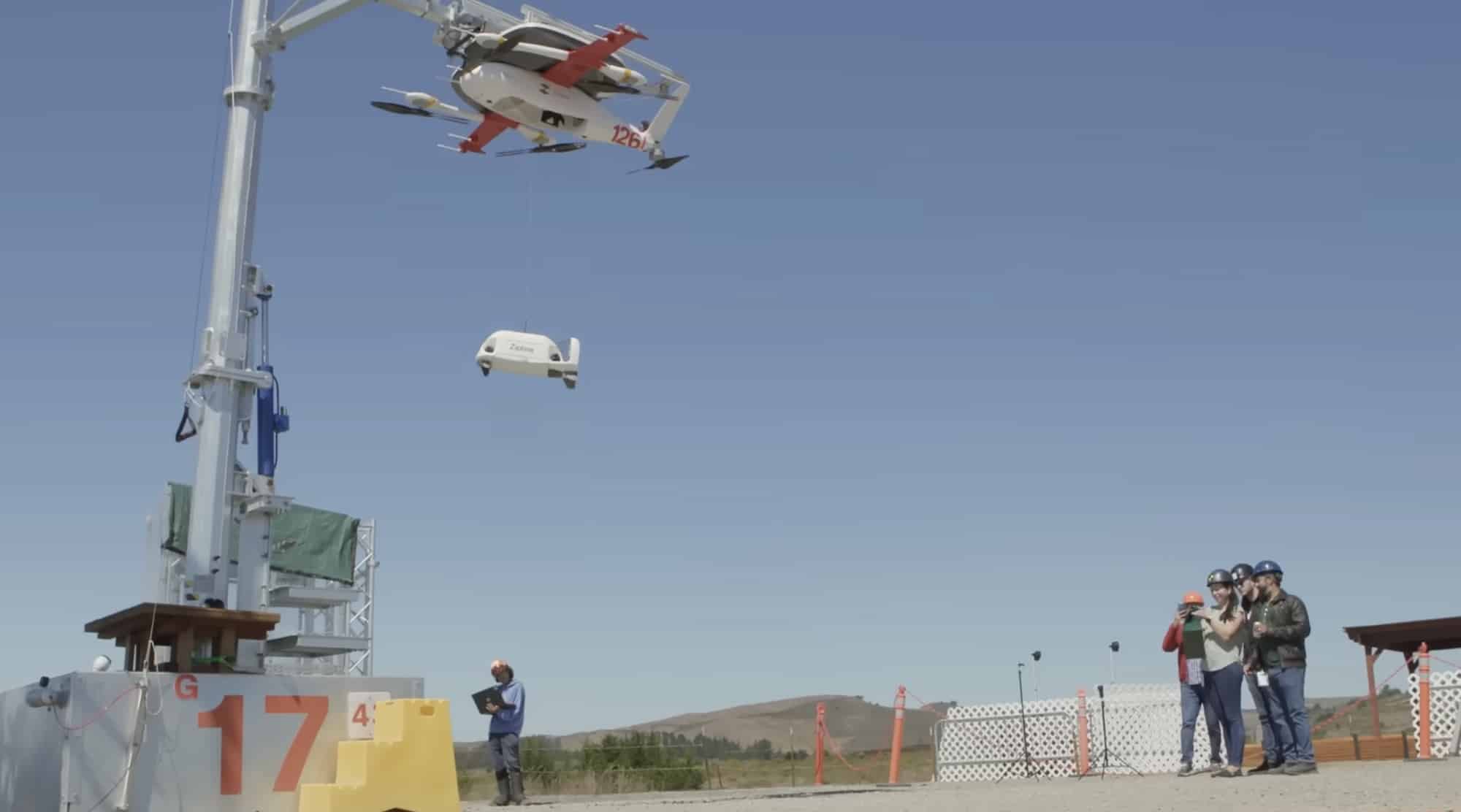 Zipline delivery droid lowering on a tether from its docking mast as observers watch.