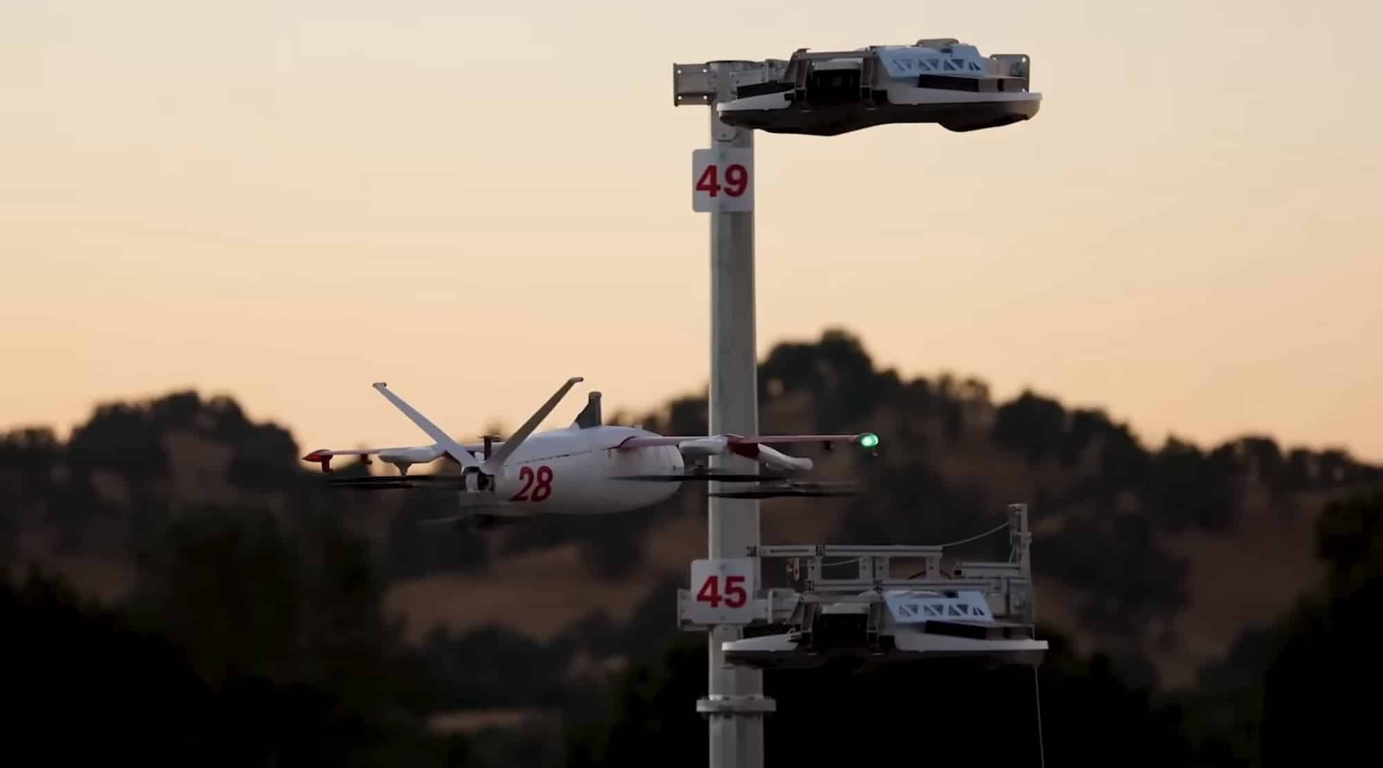 Zipline aircraft numbered 28 hovers beside its docking mast holding two numbered delivery droids.
