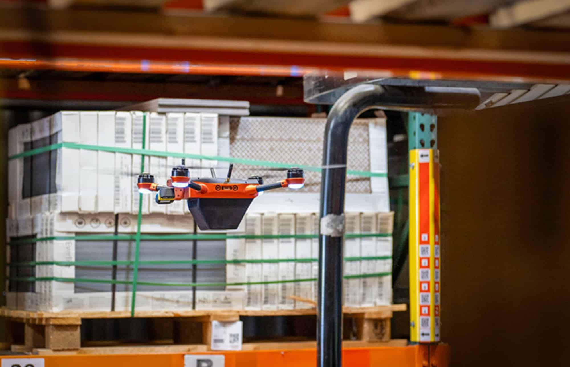 Orange Corvus inventory drone scans stacked pallets inside a narrow warehouse aisle.