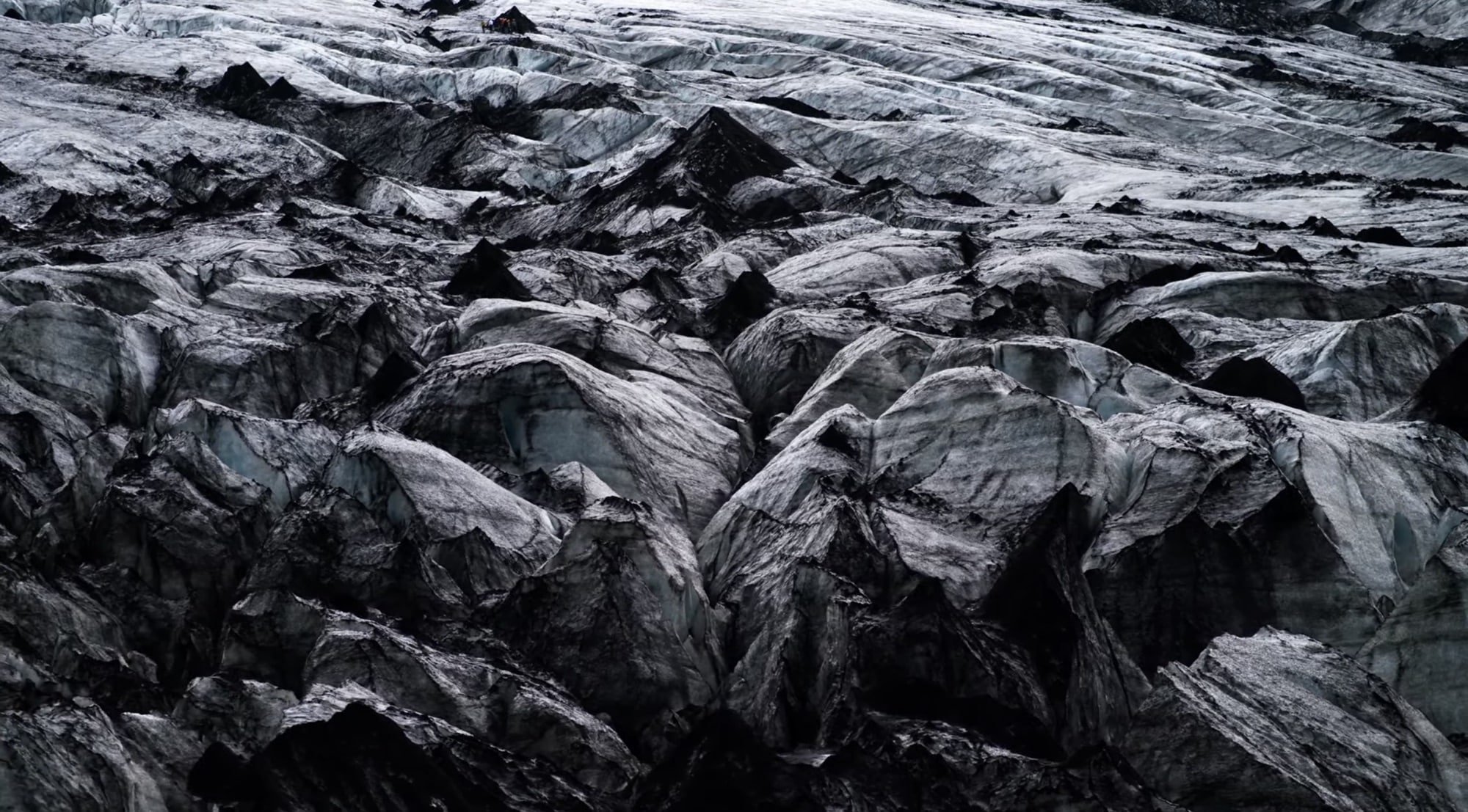 Crevassed glacier surface streaked with volcanic ash in an overhead frame.