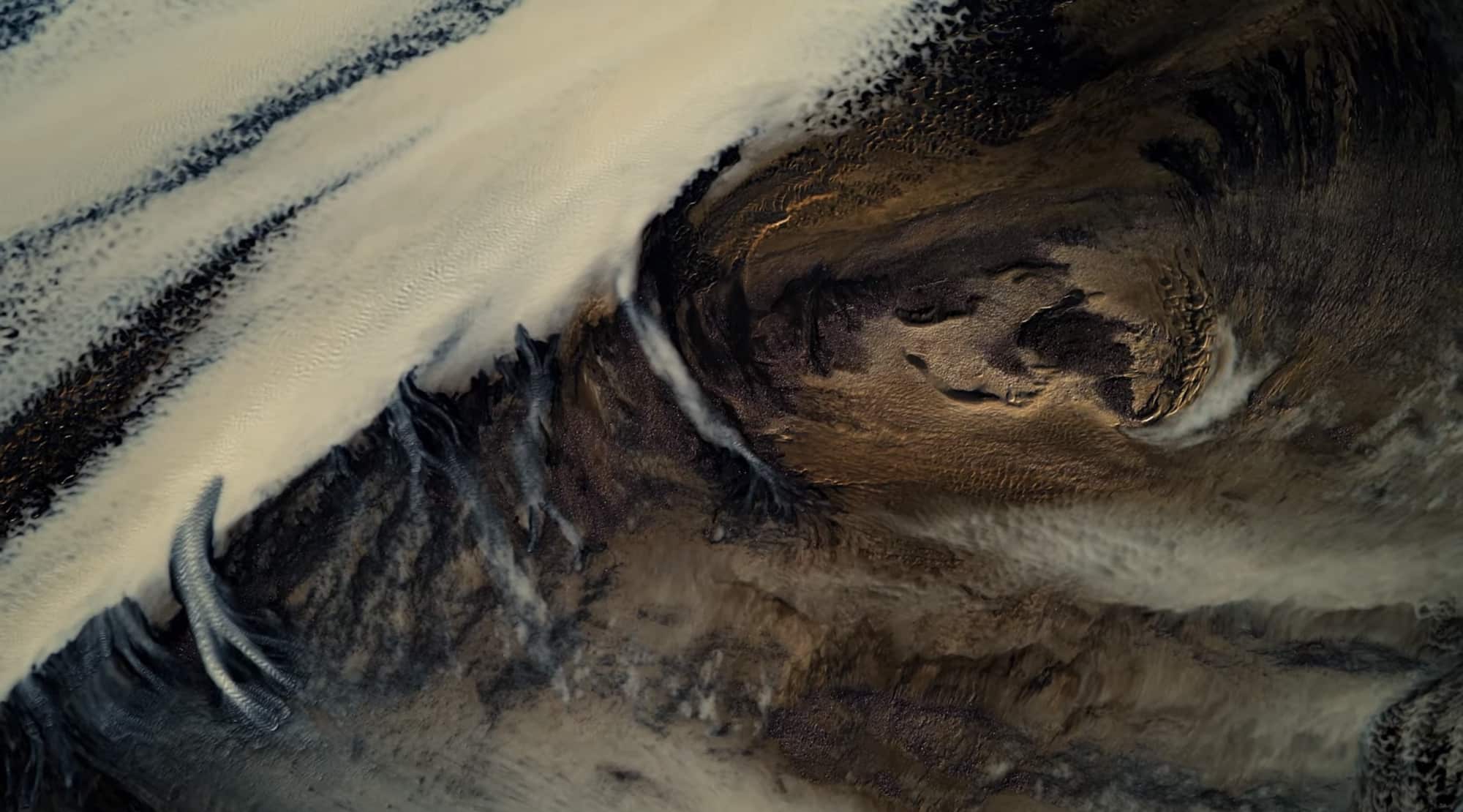 Abstract overhead frame of dark volcanic sediment swirling across pale glacial ice.