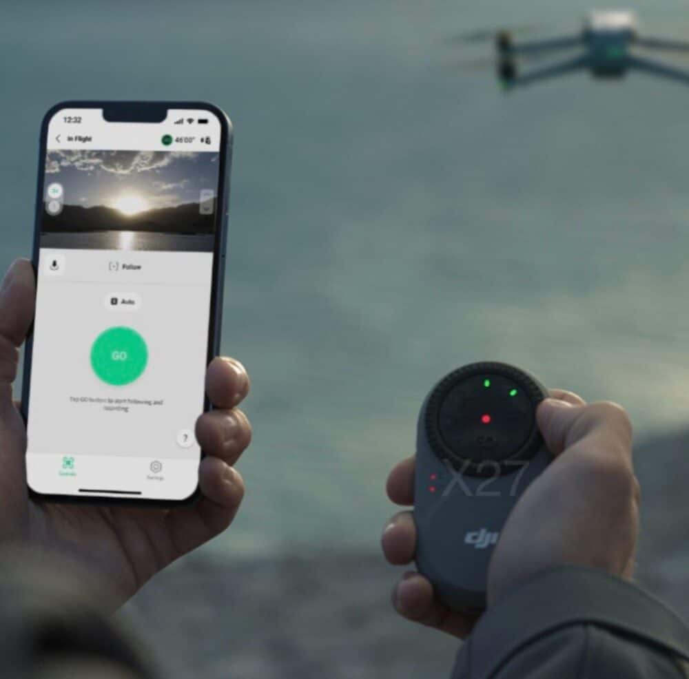 Pilot holding a phone and a small tracking beacon as a drone hovers above.